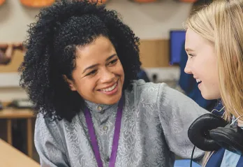 A young girl wearing headphones engages in conversation with her teacher in a classroom setting.