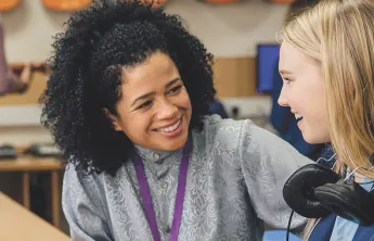 A young girl wearing headphones engages in conversation with her teacher in a classroom setting.