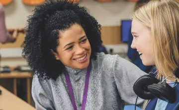 A young girl wearing headphones engages in conversation with her teacher in a classroom setting.